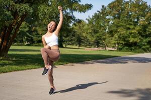 Excited young asian woman winning, finish running in park, saying yes, lifting hand up in triumph, celebrating victory or success photo