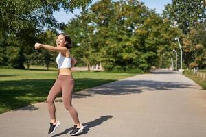 emocionado joven asiático mujer victorioso, terminar corriendo en parque, diciendo Sí, levantamiento mano arriba en triunfo, celebrando victoria o éxito foto