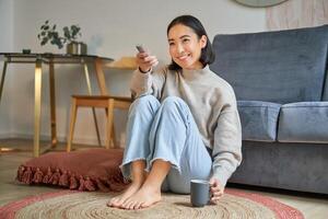 Portrait of beautiful asian girl sitting at her home and watching tv, holding remote, smiling and laughing, feeling comfort and warmth at her apartment photo