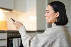 Close up portrait of smiling asian woman watching television, sitting on sofa with remote and switching chanel, looking relaxed photo