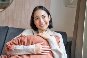 mujer y bienestar concepto. joven mujer en calentar ropa, se sienta a hogar, abrazos almohada en sofá y sonrisas complacido, piensa de algo foto