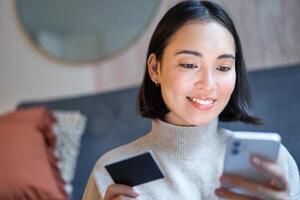 Portrait of smiling asian woman paying with credit card on her smartphone app, arrange direct debit or shopping online from mobile phone photo