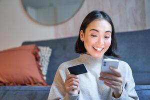 Portrait of smiling asian woman paying with credit card on her smartphone app, arrange direct debit or shopping online from mobile phone photo