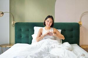 Tender asian girl shows heart sign, lying in bed, spending time in bedroom, expresses love and care photo