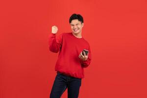 Cheerful young Asian man wearing a red sweater holding a smartphone with his fist up on red background. photo