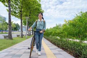 Asian woman wearing wireless headphones walking with bicycle in park. photo