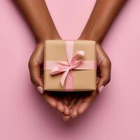 AI generated Black womans hands holding a present gift box with bow against a light pink background. Overhead view. Close up. Mothers, Women day photo