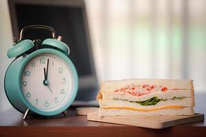 Sandwich and alarm clock displaying noon time on wooden table in office photo