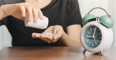 Person in a black t-shirt pouring pills into the hand with an alarm clock on the table photo