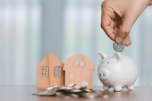 Hand saving money into a piggy bank with the wooden house and a pile of coin for financial growth and home investment concept photo