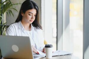 sonriente niña estudiante estudiar en línea con skype maestro, contento joven mujer aprender idioma escucha conferencia reloj seminario web escribir notas Mira a ordenador portátil sentar en cafetería, distante educación foto