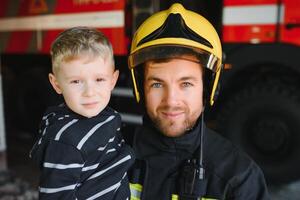 Dirty firefighter in uniform holding little saved boy standing on black background photo
