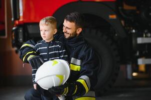 Dirty firefighter in uniform holding little saved boy standing on black background. photo