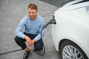 Hansome bearded guy sitting near his new modern electric car and holding plug of the charger, while car is charging at the charging station. photo