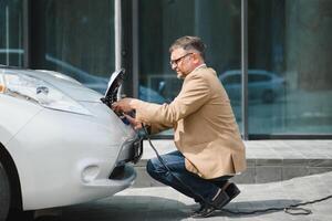 Hansome bearded man sitting near his new modern electric car and holding plug of the charger, while car is charging at the charging station photo