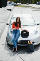 joven mujer es en pie cerca el eléctrico coche. el alquiler coche es cargando a el cargando estación para eléctrico vehículos coche intercambio. foto