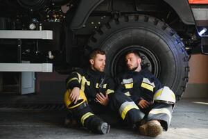 Two professional firefighters with uniforms and protective helmets posing infront of a firetruck. photo