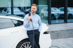 Full length portrait of young handsome bearded man in casual wear, standing at the charging station and holding a plug of the charger for an electric car. Eco electric car concept. photo