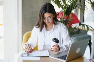 joven encantador hembra persona de libre dedicación utilizando ordenador portátil computadora para distancia trabajo mientras sentado en moderno café tienda interior, hermosa caucásico mujer trabajando en netbook durante Mañana desayuno en café bar foto