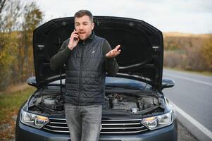 hermoso joven hombre vocación para asistencia con su coche roto abajo por el borde del camino foto