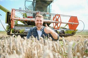 Happy farmer proudly standing in a field. Combine harvester driver going to crop rich wheat harvest. Agronomist wearing flannel shirt, looking at camera on a farmland photo