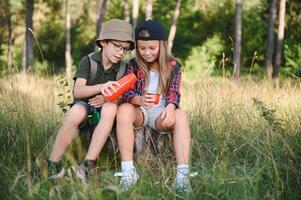 niña y chico explorar sentado y Bebiendo caliente té desde termo en el bosque foto