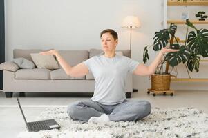 Fitness at home online. Middle aged woman in sportswear doing yoga on sports mat with laptop in living room interior with gadgets, profile, free space. photo