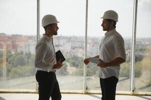 A front view of two smart architects with white helmets reviewing blueprints at a construction site on a bright sunny day photo