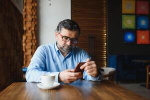 contento empresario sentado en restaurante y esperando para almuerzo. él es utilizando inteligente teléfono y hablando con alguien. negocio personas mayores estilo de vida concepto. foto