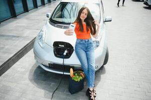 Young woman is standing near the electric car. The rental car is charging at the charging station for electric vehicles. Car sharing. photo