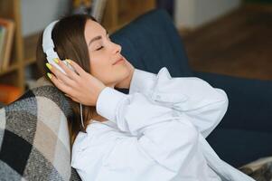 Portrait of attractive girl 20s wearing headphones using smartphone and listening to music while sitting on sofa at home photo