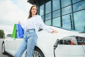 Happy young adult woman smiling wide, looking away, charging automobile battery from small public station, standing near electric car. photo