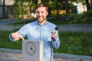 Portrait of a young man standing with charging cable near the charging station. Concept of fast home car chargers photo