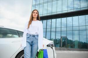 contento joven mujer en pie en ciudad estacionamiento cerca eléctrico auto, cargando automóvil batería desde pequeño ciudad estación, participación compras bolsas. foto