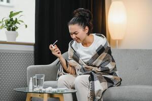 sick young woman in warm clothes spraying throat while sitting on couch at home. photo