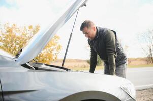 hombre reparando un roto coche por el la carretera. hombre teniendo problema con su roto coche en el autopista borde del camino. hombre mirando debajo el coche capucha. coche rompe abajo en el autopista. borde del camino asistencia concepto. foto