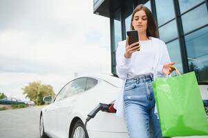 contento joven adulto mujer sonriente ancho, mirando lejos, cargando automóvil batería desde pequeño público estación, en pie cerca eléctrico coche. foto