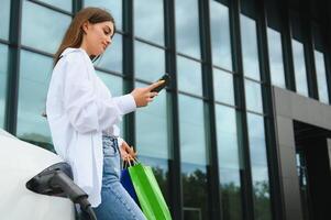 Happy young woman standing on city parking near electric car, charging automobile battery from small city station, holding shopping bags. photo