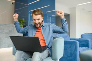 foto de joven emocionado hombre contento positivo sonrisa celebrar ganar victoria éxito puños manos sentar sofá hogar remoto trabajar.