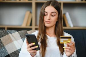 contento adolescente haciendo compras en línea sentado en sofá a hogar. joven mujer insertar crédito tarjeta número en sitio web con su inteligente teléfono. foto