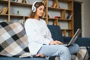 sonriente atractivo joven mujer sentado en sofá utilizando ordenador portátil comunicado trabajando en línea a hogar, contento adolescente niña mecanografía en computadora, disfrutando escritura Blog o chateando con amigos en social red foto