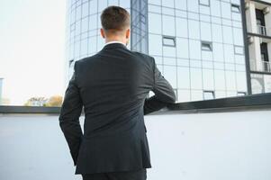 moderno empresario. confidente joven hombre en lleno traje y mirando lejos mientras en pie al aire libre con paisaje urbano en el antecedentes foto