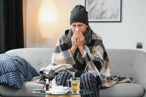 Sick sad man sits on couch at home suffers from runny nose flu disease coronavirus pandemic covid epidemic sneezes. Unwell guy feeling bad fever virus illness symptoms indoor. photo