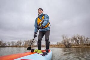 senior male paddler is paddling a stand up paddleboard in cold winter weather on a calm lake photo
