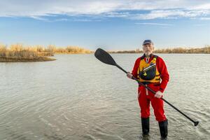 environmental portrait of a senior male paddler in a drysuit and life jacket holding stand up paddle on a shore of a lake photo