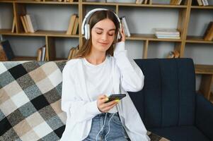 Portrait of attractive girl 20s wearing headphones using smartphone and listening to music while sitting on sofa at home photo