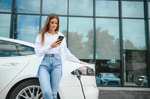 teléfono en manos. mujer en el eléctrico carros cargar estación a tiempo de día. marca nuevo vehículo. foto