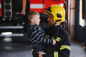 Dirty firefighter in uniform holding little saved boy standing on black background. photo