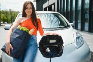 joven mujer es en pie cerca el eléctrico coche. el alquiler coche es cargando a el cargando estación para eléctrico vehículos coche intercambio. foto