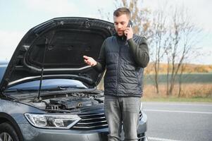 hermoso joven hombre vocación para asistencia con su coche roto abajo por el borde del camino foto